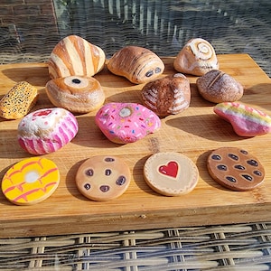 May include: A collection of thirteen smooth stones painted to resemble various baked goods, including a pink frosted doughnut, chocolate chip cookies, a heart-decorated cookie, a yellow glazed pastry, a cupcake, and several bread-like rolls.