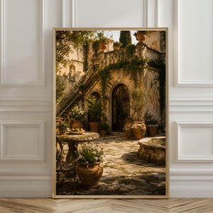 Op de afbeelding: Een ingelijste wandkunstprint met een zonnige mediterrane binnenplaats met stenen architectuur, een grote trap, klimop en terracotta potten gevuld met groen en bloemen op een terras van verweerde stenen.