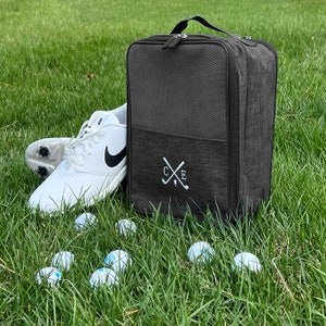 May include: A grey shoe bag with a mesh top and a zip closure. The bag has a white embroidered logo with the letters "C.E." and two crossed golf clubs. A white golf shoe is next to the bag, and there are several white golf balls scattered on the green grass.