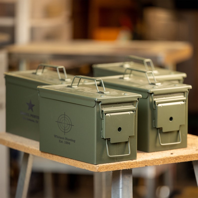 Peut inclure: Trois bo&icirc;tes &agrave; munitions en m&eacute;tal vert olive avec couvercles &agrave; charni&egrave;re et poign&eacute;es de transport. Les bo&icirc;tes ont une forme rectangulaire et sont &eacute;tiquet&eacute;es avec du texte blanc et un graphique de cible. Le texte indique "Whitson Hunting Est. 1994" et "HOUSTON, TX".