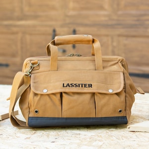May include: A tan canvas tool bag with the name "LASSITER" printed in black. The bag has a top handle, a shoulder strap, and a front pocket with two silver button closures. The bottom of the bag is a dark gray color.