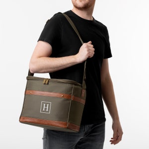 K&ouml;nnte beinhalten: Eine gr&uuml;ne K&uuml;hltasche aus Canvas mit braunen Lederelementen und einem Rei&szlig;verschluss. Die Tasche hat einen Schultergurt und ein Monogramm "H" auf der Vorderseite.
