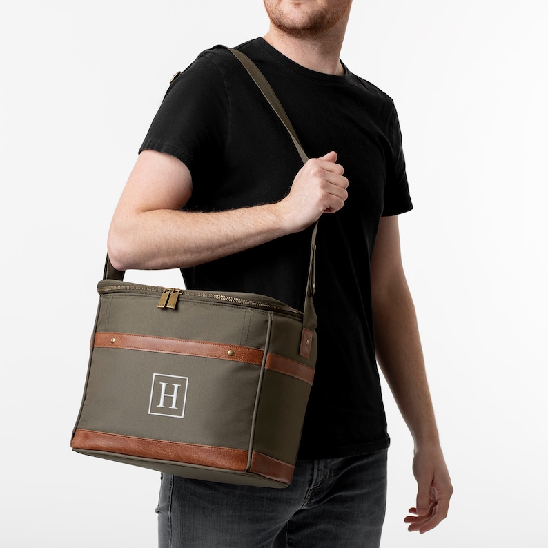K&ouml;nnte beinhalten: Eine gr&uuml;ne K&uuml;hltasche aus Canvas mit braunen Lederelementen und einem Rei&szlig;verschluss. Die Tasche hat einen Schultergurt und ein Monogramm "H" auf der Vorderseite.