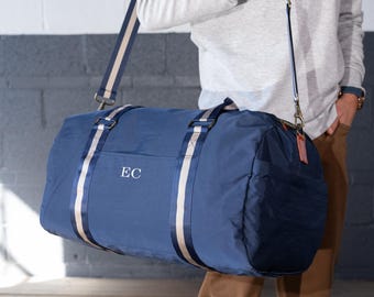May include: Navy blue duffel bag with cream and navy stripes. The bag has a shoulder strap and a side pocket. The initials "EC" are embroidered on the front. The bag is held by a person.