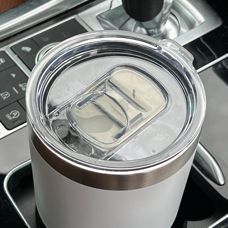 Puede incluir: Primer plano de un vaso t&eacute;rmico blanco con tapa de pl&aacute;stico transparente. La tapa tiene un mecanismo deslizante para abrir y cerrar la boquilla. El vaso tiene una banda de acero inoxidable en la parte superior e inferior.