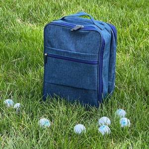 May include: A blue canvas backpack with a zipper closure and a dark blue trim. The backpack is sitting on a green grassy surface with several white balls scattered around it.