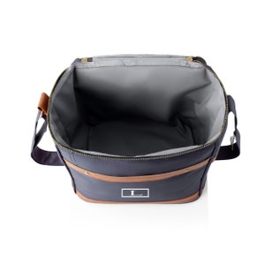 K&ouml;nnte beinhalten: Eine isolierte K&uuml;hltasche in Marineblau mit braunem Lederbesatz und Rei&szlig;verschluss. Die Tasche hat ein wei&szlig;es Monogramm "L" auf der Vorderseite.