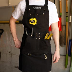 May include: Black work apron with multiple pockets, a yellow tape measure, and tools. The apron has the name "HAWKINS" printed on the top. The apron has a metal hook and a metal ring on the bottom.