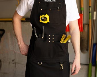 Puede incluir: Delantal de trabajo negro con múltiples bolsillos, un flexómetro amarillo y herramientas. El delantal tiene el nombre "HAWKINS" impreso en la parte superior. El delantal tiene un gancho y un anillo de metal en la parte inferior.
