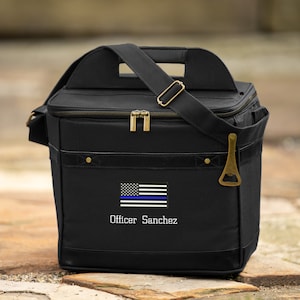 May include: Black canvas cooler bag with a thin blue line American flag and the text "Officer Sanchez". The bag has a zipper closure, a top handle, and a shoulder strap. It also has a built-in bottle opener.