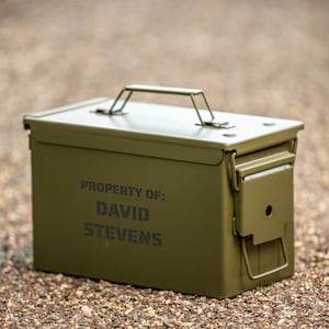 Peut inclure: Une bo&icirc;te &agrave; munitions en m&eacute;tal vert olive avec un couvercle &agrave; charni&egrave;re et une poign&eacute;e en m&eacute;tal. Le texte "PROPERTY OF: DAVID STEVENS" est imprim&eacute; sur le devant. La bo&icirc;te est pos&eacute;e sur une surface de petits cailloux.