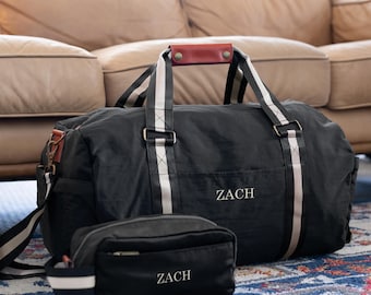 May include: Black duffel bag with white stripes and a brown leather handle. The bag has a zippered pocket on the front and the name "ZACH" embroidered on it. A smaller black toiletry bag with the same name is in front of the duffel bag.