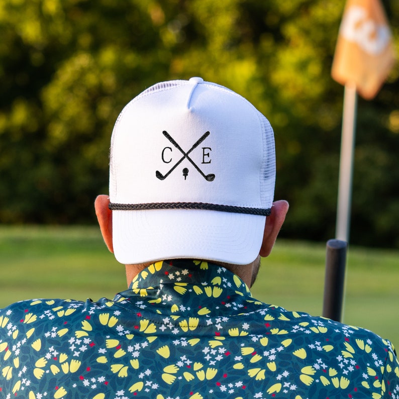 Puede incluir: Gorra de camionero blanca con un dise&ntilde;o bordado en negro de palos de golf cruzados y las letras "C E".