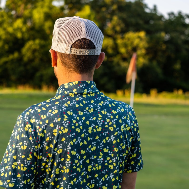Puede incluir: Una persona con una gorra de b&eacute;isbol blanca y gris y una camisa con estampado azul y amarillo. La persona est&aacute; de pie en un campo de hierba verde.