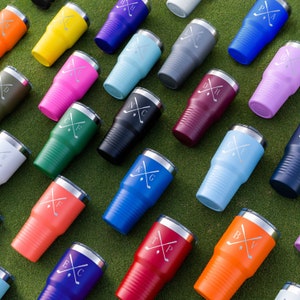 Puede incluir: Una colecci&oacute;n de vasos aislados en varios colores, incluyendo amarillo, rosa y azul, dispuestos sobre una superficie verde. Cada vaso presenta una tapa plateada y un dise&ntilde;o de palos de golf cruzados con iniciales. Los vasos est&aacute;n dise&ntilde;ados para mantener las bebidas fr&iacute;as o calientes.