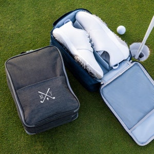 May include: A dark grey shoe bag with a white embroidered monogram "MY" and crossed golf clubs. A blue shoe bag with a white lining is open and contains two white golf shoes.