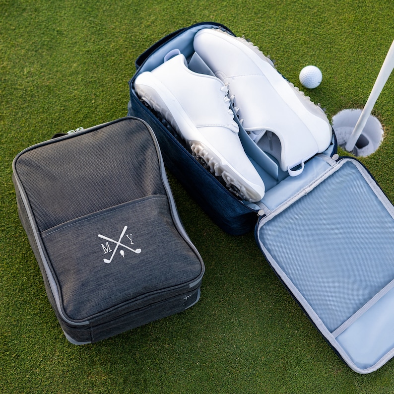 May include: A dark grey shoe bag with a white embroidered monogram "MY" and crossed golf clubs. A blue shoe bag with a white lining is open and contains two white golf shoes.