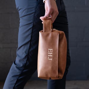 May include: A brown leather travel toiletry bag with a zipper closure and a top handle. The bag has the initials "EHJ" embroidered on the front in gold thread.