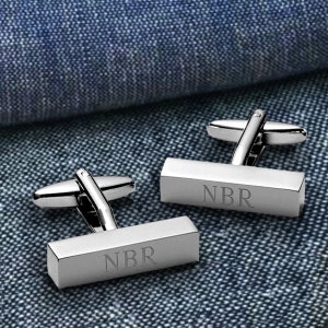 May include: A pair of silver cufflinks with a rectangular shape. The cufflinks are engraved with the initials "NBR".
