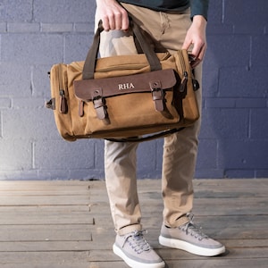 May include: A brown canvas duffel bag with a leather flap and straps. The bag has a top handle and a shoulder strap. The leather flap is embossed with the initials "RHA". The bag is held by a person wearing khaki pants and gray sneakers.