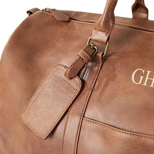 May include: A brown leather duffel bag with a luggage tag and the initials "GH" embroidered on the front.