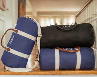 May include: Three personalized duffel bags in navy blue and black. The navy bags have white stripes and tan handles, with the names "James" and "Nick" embroidered. The black bag has the initials "EHV". All bags have a shoulder strap.
