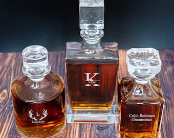 May include: Three clear glass decanters with stoppers, filled with amber liquid. The decanters are engraved with the letters "K", "L", and the text "Colin Robinson Groomman".