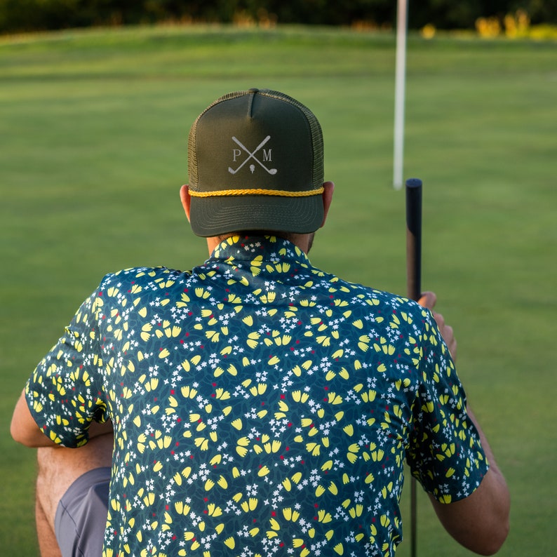 Puede incluir: Una persona lleva una camisa con estampado verde y amarillo con una gorra de b&eacute;isbol verde oscuro que dice "P.M." y un dise&ntilde;o de palo de golf cruzado blanco. La persona sostiene un palo de golf.
