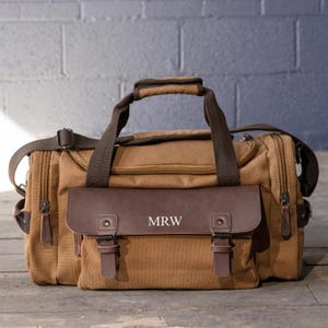 May include: A brown canvas duffel bag with a brown leather flap and accents. The bag has a top handle, a shoulder strap, and the initials "MRW" are visible on the flap. The bag has multiple zippered compartments.