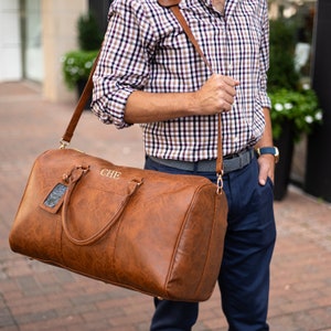 May include: A brown leather duffel bag with a gold strap and a gold name tag. The bag is monogrammed with the letters "CHE".