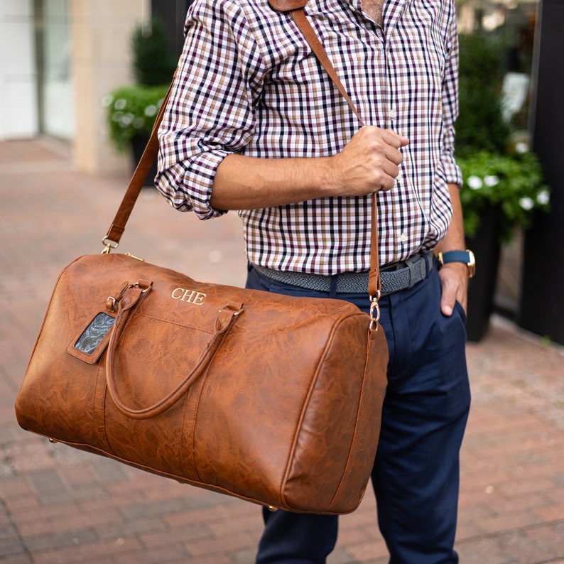 May include: A brown leather duffel bag with a gold strap and a gold name tag. The bag is monogrammed with the letters "CHE".
