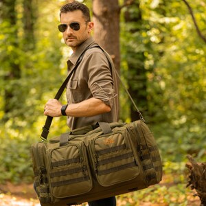 May include: An olive green duffel bag with multiple pockets and straps is carried over the shoulder. The bag has the initials "GAL" embroidered in orange. The person carrying the bag is wearing sunglasses and a long-sleeved shirt.