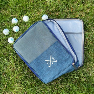 May include: A blue golf ball bag with a zipper closure and a monogrammed design featuring the initials "C.B." and crossed golf clubs. The bag is open and lying on a bed of green grass with several golf balls scattered around it.