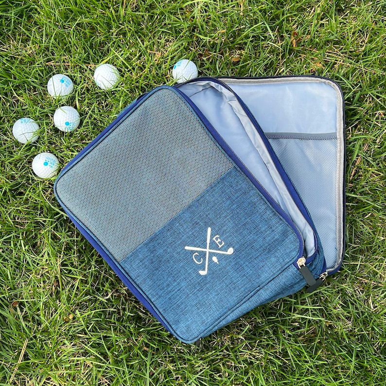 May include: A blue golf ball bag with a zipper closure and a monogrammed design featuring the initials "C.B." and crossed golf clubs. The bag is open and lying on a bed of green grass with several golf balls scattered around it.