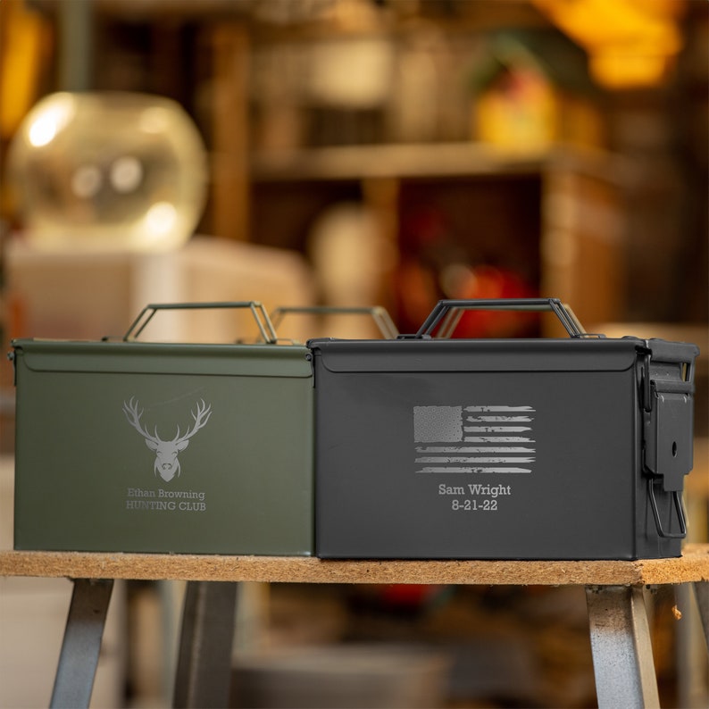 Peut inclure: Deux bo&icirc;tes &agrave; munitions en m&eacute;tal. L'une est vert olive avec un graphique de t&ecirc;te de cerf et le texte "Ethan Browning HUNTING CLUB". L'autre est noire avec un drapeau am&eacute;ricain et le texte "Sam Wright 8-21-22". Les deux ont des poign&eacute;es.