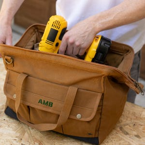 May include: A brown canvas tool bag with a zipper closure, two handles, and a front pocket. The bag has the initials "AMB" embroidered in green. A yellow and black drill is being placed inside the bag.
