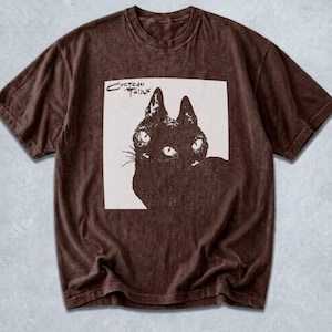 May include: A brown short-sleeve t-shirt with a washed-out texture. The center features a square graphic with a cream background displaying a black illustration of a cat with multiple eyes and the text Cocteau Twins printed in the upper left corner.