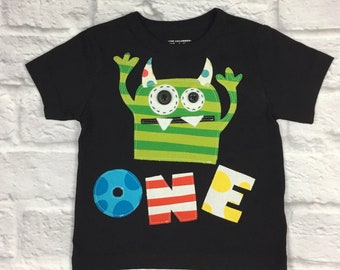 May include: Black short-sleeved t-shirt with a green monster graphic and the word "ONE" in colorful letters. The monster has a zipper mouth, button eyes, and green and white stripes.