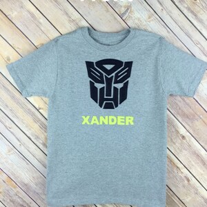 May include: Gray t-shirt with a black Transformers logo and the name "XANDER" in yellow lettering.