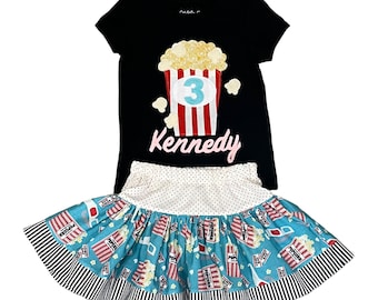 May include: Black t-shirt with a red and white popcorn bucket applique with a blue number 3 and the name Kennedy in pink. A blue and white polka dot skirt with a popcorn and 3D glasses print.