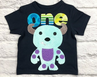May include: A navy blue short-sleeved t-shirt with a felt applique of a blue monster with purple accents. The monster has large eyes and a toothy grin. The word "one" is sewn on the shirt in colorful fabric.