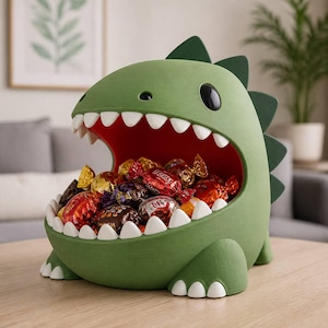 May include: A green dinosaur-shaped bowl with a wide-open mouth filled with assorted wrapped candies. The creature features a smooth matte finish, white pointed teeth, a deep red interior, and dark green dorsal spikes along its back.