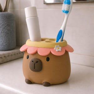 Pode incluir: Um suporte para escovas de dentes em forma de capivara castanha com rebordo superior amarelo e uma borda de pétalas cor-de-rosa. O suporte apresenta uma pequena flor branca, um nariz castanho escuro e dois pequenos pés castanhos. Contém um tubo.