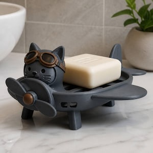May include: A dark gray soap dish shaped like a small airplane with a cat pilot wearing brown goggles. The cat is positioned at the front behind a propeller, and a rectangular off-white bar of soap rests in the center of the airplane body.