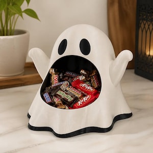 May include: A white ceramic ghost-shaped bowl with a wide, open mouth serving as a container. The ghost has two black oval eyes and a black base. The interior of the mouth is filled with various individually wrapped chocolate candy bars.