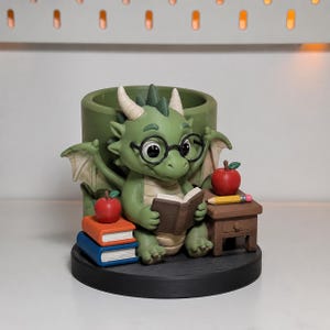 Pode incluir: Uma estatueta de dragão verde com chifres brancos e óculos redondos pretos, segurando um livro aberto. O dragão senta-se numa base preta em frente a um recipiente cilíndrico verde, com uma pilha de livros laranja e azuis, uma maçã vermelha.