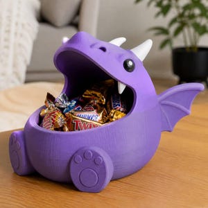 May include: A purple dragon-shaped bowl with a wide-open mouth filled with assorted sweets. The creature features white horns, a single visible white tooth, and small rounded feet. The surface of the bowl shows a textured 3D printed finish.