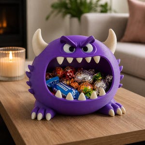 May include: A purple monster-shaped bowl with a wide open mouth filled with various wrapped candies. The creature features two large white horns, sharp white teeth lining the mouth, and a pair of angry-looking eyes with thick purple brows.