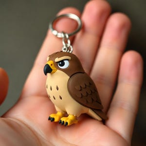 May include: A keychain featuring a small, stylized falcon figurine with brown and tan plumage, a yellow beak with a black tip, and yellow feet with black talons. The bird has an intense expression with white eyes and black pupils, attached to a silver metal ring.