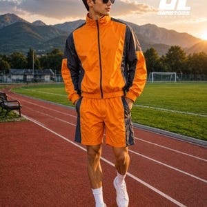 May include: A matching two-piece athletic set consisting of a long-sleeve track jacket with a full-length zipper and matching shorts. The set features a vibrant orange base color with dark charcoal gray side panels and black trim details.